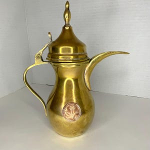 May include: A vintage brass coffee pot with a long spout, curved handle, and a decorative finial on the lid. The pot features a copper emblem with a palm tree design. The overall design is ornate and traditional.
