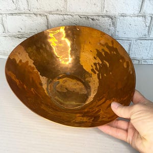 May include: A large, round, polished copper bowl with a distinct hammered texture. The interior surface features a warm, metallic orange-brown finish with visible light reflections and subtle indentations across the entire curved metal body.