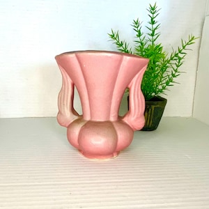 May include: A vintage pink ceramic vase with a unique, fluted design. The vase features two curved handles and a bulbous base. The vase is approximately 7 inches tall and is a soft, muted pink color.