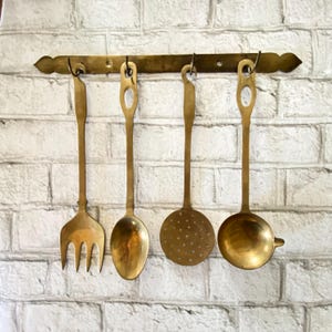 May include: A set of four antique brass kitchen utensils hanging on a brass bar. The set includes a fork, spoon, slotted spoon, and ladle. The utensils are suspended against a white brick wall.