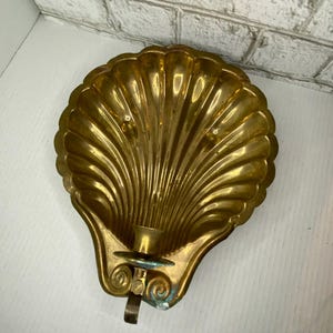 May include: A vintage brass seashell-shaped candle holder. The candle holder has a detailed, ridged design and a small cup to hold a single candle. The holder has a decorative bracket and a small, round base.