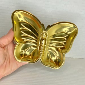 May include: A gold-colored butterfly-shaped dish. The dish has detailed wing patterns and a textured body. The wings are curved, and the dish is held by a hand. The dish is made of metal and has a shiny finish.