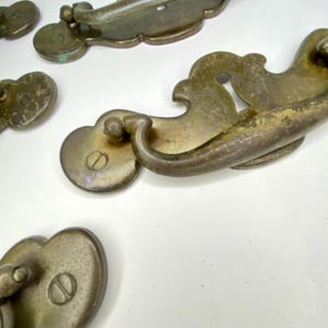 May include: Close-up of several antique brass drawer pulls. The pulls have a curved design with a keyhole detail and a decorative backplate. The brass has a tarnished, aged appearance, suggesting vintage or reclaimed hardware.