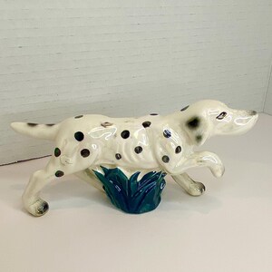 May include: A ceramic figurine of a Dalmatian dog running through a patch of green grass. The dog is white with black spots and has a black nose and ears.