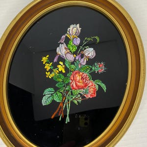 May include: An oval wall art piece featuring a gold-toned frame surrounding a black background. A hand-painted floral bouquet is centered on the black surface, showcasing red roses, purple irises, and small yellow flowers with green leaves.