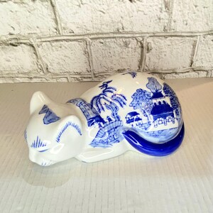 May include: A white ceramic cat figurine with blue transferware detailing. The cat is in a resting pose, with a blue tail and blue landscape scenes decorating its body. The figurine is a decorative item.
