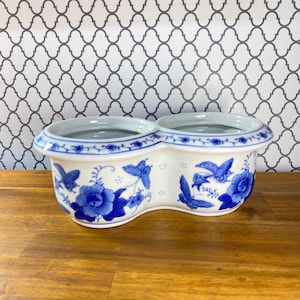 May include: A white ceramic double planter featuring a blue floral and butterfly pattern. The vessel has two circular openings with a wide, rounded rim decorated with a delicate blue vine motif encircling the top edge of the container.