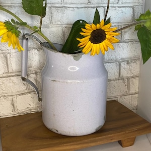 May include: A light gray enameled metal pitcher with a handle and visible wear, containing two yellow sunflowers with green stems and leaves. The pitcher features a cylindrical body with a wide rim and shows signs of chipped enamel finish.