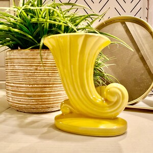May include: A yellow ceramic vase with a unique, curved design. The vase has a wide opening and a narrow base. It is a decorative piece that would add a touch of color to any home.