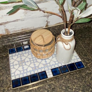 May include: A gold metal tray with a blue and white tile design featuring a heart pattern. The tray has a wooden handle and is holding a white ceramic vase with a plant and a glass jar with a wooden lid.