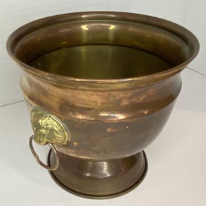 May include: A vintage copper planter with a brass lion's head detail. The planter has a wide, rounded bowl shape with a pedestal base. The copper has a warm, aged patina, and the brass lion's head adds a decorative touch.
