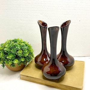 May include: Three brown glass vases with a glossy finish. The vases have a teardrop shape with a slender neck and a flared opening. They are arranged on a vintage book, with a small green plant in a brown pot to the left.