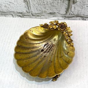 May include: A decorative, antique-style seashell-shaped dish with a gold finish. The dish features a scalloped edge and is adorned with gold-colored roses and leaves. The dish is likely made of metal.
