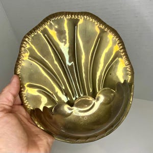 May include: A vintage brass shell-shaped dish with a scalloped edge. The dish has a detailed, textured surface with a glossy finish. The design features radiating lines and a curved, three-dimensional base, creating a decorative and functional piece.