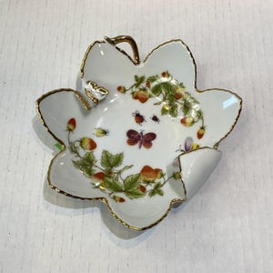 May include: White leaf-shaped porcelain dish with a gold rim. The dish is decorated with strawberries, green leaves, and small insects. A gold butterfly is on the side. A decorative piece for serving or display.