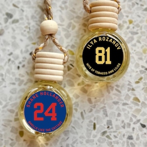 May include: Two small glass bottles with wooden caps and hanging cords. One bottle has a blue label with the number 24 and the text "SHANE HOLLANDER SMELLS LIKE THE COTTAGE". The other has a black label with the number 81 and the text "ILYA ROZANOV HITS OF TOBACCO AND LILIES".