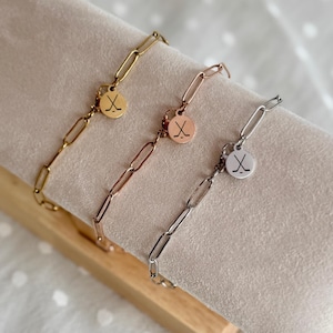 May include: Three link chain bracelets in gold, rose gold, and silver. Each bracelet has a small, round charm with a crossed golf club design. The bracelets are displayed on a beige velvet surface.