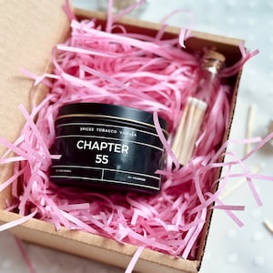 Pode incluir: Um frasco de vela preto com o texto "SPICES TOBACCO VANILLA CHAPTER 55" no rótulo. A vela está em uma caixa de papelão marrom com papel picado rosa.