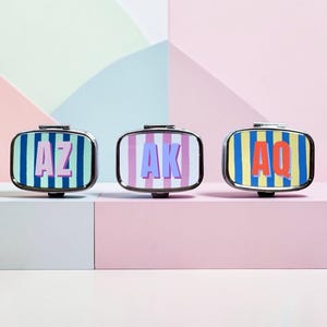 Puede incluir: Tres pastilleros rectangulares con marco plateado y diseños de rayas coloridas. Cada caja presenta una inicial de dos letras diferente: "AZ" en morado, "AK" en lavanda y "AQ" en rojo. El fondo es un patrón geométrico pastel.