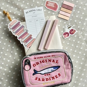 May include: A pink and clear plastic pouch with a zipper closure. The pouch has an embroidered design of a can of sardines with the text "Original Sardines" and "In Pure Olive Oil".