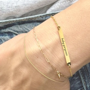 May include: A gold bar bracelet with the inscription "baby gorgeous" on a woman's wrist. The bracelet is layered with two other gold chain bracelets.