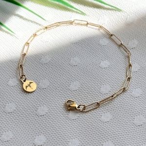 Puede incluir: Un brazalete de cadena dorada con un pequeño dije con un diseño de pájaro. El brazalete está sobre un fondo blanco con una sombra de hoja verde.