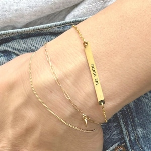 May include: Gold-toned bracelets stacked on a wrist, including a delicate chain, a paperclip chain, and a bar bracelet engraved with "name 'em". The bracelets are worn with a gray t-shirt and blue jeans.