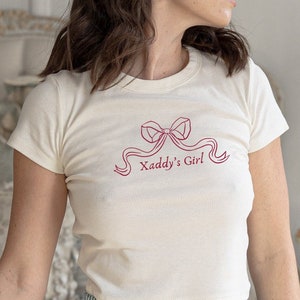 Puede incluir: Top corto color crema con el texto "Xaddy's Girl" en un diseño de lazo rosa. Combinado con shorts a rayas azules y blancas. El conjunto es informal y adecuado para ropa de dormir.