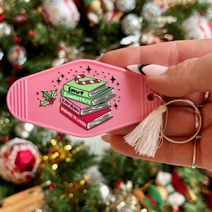 Może przedstawiać: Różowy brelok w kształcie znaku motelu z stosami książek, na których napisano "Smut", "Romantasy", "Slow Burn" i "Enemies to Lovers".