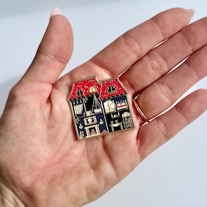 May include: A gold-toned metal enamel pin shaped like a two-story house with a red roof. The interior rooms feature black walls and detailed illustrations of furniture, a fireplace, a bed, and various household items in white and gold outlines.