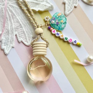Peut inclure: Un flacon diffuseur d'huiles essentielles en verre transparent avec un bouchon en bois et un cordon beige. Le diffuseur est orné d'un charme en forme de coquillage et d'une étiquette nominative perlée qui dit "AMANDA".