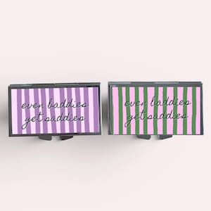 Op de afbeelding: Twee rechthoekige metalen pillendoosjes. E&eacute;n heeft een paars en wit gestreept patroon, de andere een roze en groen gestreept patroon. Beide doosjes hebben de tekst "even baddies get saddies" in cursief.
