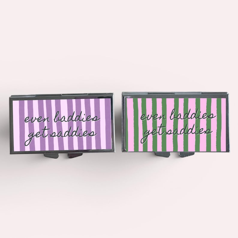 Op de afbeelding: Twee rechthoekige metalen pillendoosjes. E&eacute;n heeft een paars en wit gestreept patroon, de andere een roze en groen gestreept patroon. Beide doosjes hebben de tekst "even baddies get saddies" in cursief.