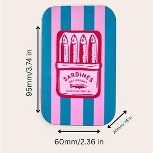 May include: A rectangular metal tin featuring vertical pink and teal stripes. The center displays a graphic of four sardines in a box with the text SARDINES NET DREAMS IN LOVE BY THE SEA. Dimensions are 3.74 inches by 2.36 inches by 0.78 inches.