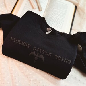 Puede incluir: Sudadera negra de cuello redondo con el texto "VIOLENT LITTLE THING" y un gr&aacute;fico de murci&eacute;lago. La sudadera est&aacute; doblada sobre una superficie blanca con lunares, con un libro abierto y dos objetos rectangulares en el fondo.