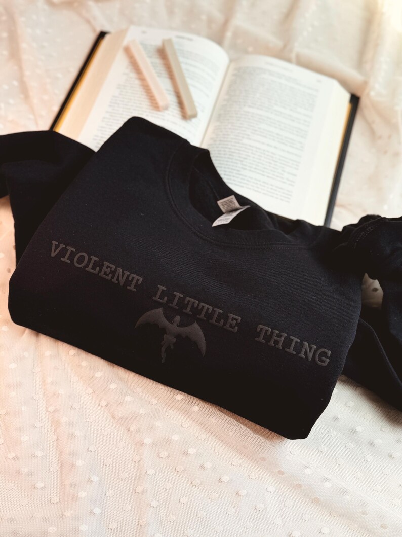 Puede incluir: Sudadera negra de cuello redondo con el texto "VIOLENT LITTLE THING" y un gr&aacute;fico de murci&eacute;lago. La sudadera est&aacute; doblada sobre una superficie blanca con lunares, con un libro abierto y dos objetos rectangulares en el fondo.