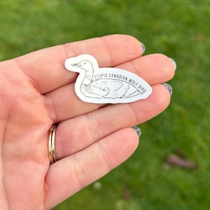 K&ouml;nnte beinhalten: Ein wei&szlig;er Aufkleber mit einer schwarzen Umrisszeichnung eines Vogels und dem Text "STUPID CANADIAN WOLF BIRD". Der Aufkleber wird in einer Hand mit einem goldenen Ring gehalten. Der Hintergrund ist verschwommenes gr&uuml;nes Gras.