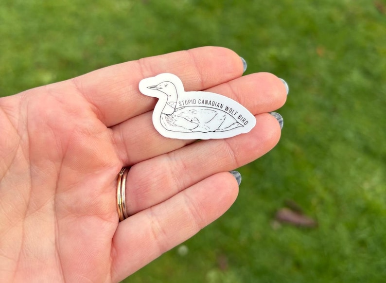 K&ouml;nnte beinhalten: Ein wei&szlig;er Aufkleber mit einer schwarzen Umrisszeichnung eines Vogels und dem Text "STUPID CANADIAN WOLF BIRD". Der Aufkleber wird in einer Hand mit einem goldenen Ring gehalten. Der Hintergrund ist verschwommenes gr&uuml;nes Gras.