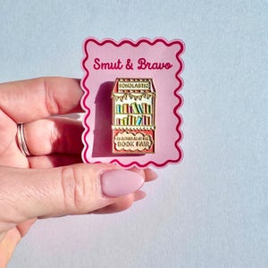 May include: A metal enamel pin shaped like a book fair display case with red and white striped accents, featuring the text Scholastic and I'd rather be at the book fair. The pin is attached to a pink scalloped backing card labeled Smut & Bravo.