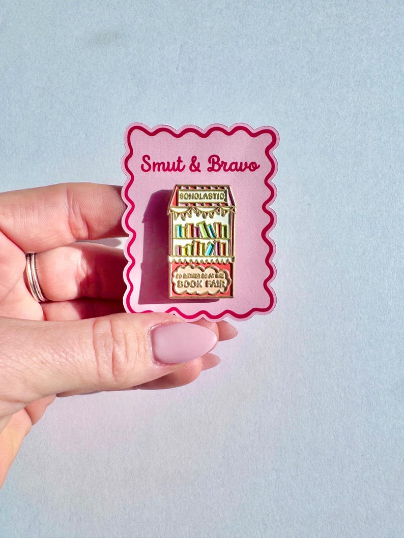May include: A metal enamel pin shaped like a book fair display case with red and white striped accents, featuring the text Scholastic and I'd rather be at the book fair. The pin is attached to a pink scalloped backing card labeled Smut & Bravo.