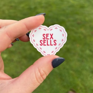 K&ouml;nnte beinhalten: Ein wei&szlig;er herzf&ouml;rmiger Aufkleber mit rotem, gewelltem Rand und den Worten "SEX SELLS" in roten Blockbuchstaben. Der Aufkleber wird vor einem verschwommenen gr&uuml;nen Hintergrund gehalten.