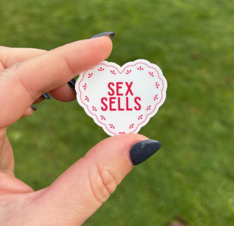 K&ouml;nnte beinhalten: Ein wei&szlig;er herzf&ouml;rmiger Aufkleber mit rotem, gewelltem Rand und den Worten "SEX SELLS" in roten Blockbuchstaben. Der Aufkleber wird vor einem verschwommenen gr&uuml;nen Hintergrund gehalten.