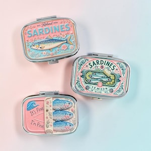 Könnte beinhalten: Drei rechteckige Metalldosen mit Klappdeckeln, die jeweils bunte Illustrationen zum Thema Sardinen zeigen. Eine Dose zeigt einen einzelnen Fisch, eine weitere mehrere Fische mit Oliven und die dritte drei Fische mit dem Text It's Fine I'm Fine.