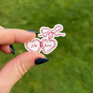 K&ouml;nnte beinhalten: Ein wei&szlig;er Aufkleber mit zwei verbundenen Herzen, die jeweils rot umrandet sind. Die Namen "Lily" und "Jane" sind in roter Schrift in den Herzen geschrieben. Eine rote Schleife ziert die Oberseite. Der Aufkleber wird vor einem gr&uuml;nen Hintergrund gehalten.