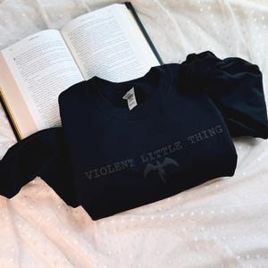 Puede incluir: Una sudadera negra con el texto "VIOLENT LITTLE THING" y un dise&ntilde;o de criatura alada. La sudadera est&aacute; doblada sobre una superficie blanca texturizada junto a un libro abierto.