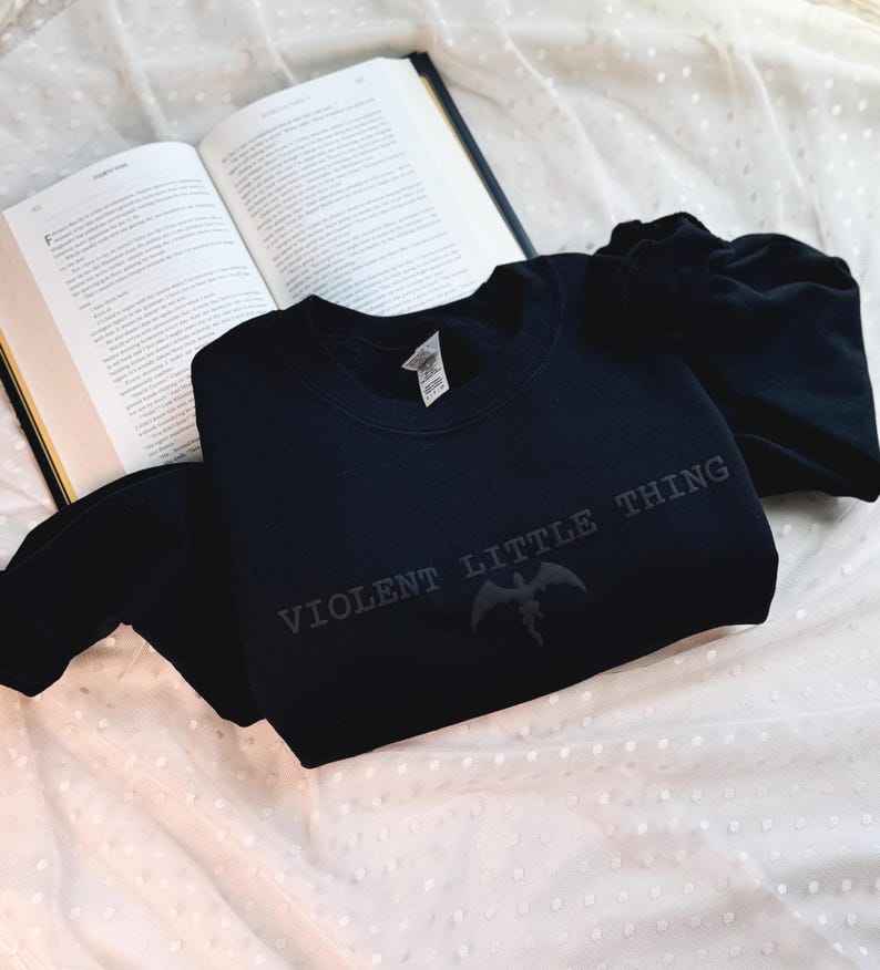 Puede incluir: Una sudadera negra con el texto "VIOLENT LITTLE THING" y un dise&ntilde;o de criatura alada. La sudadera est&aacute; doblada sobre una superficie blanca texturizada junto a un libro abierto.