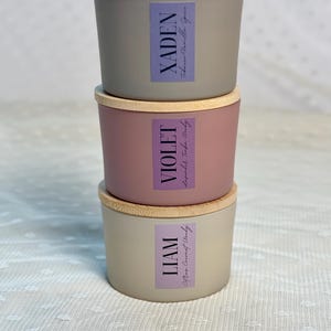 Może przedstawiać: Trzy ułożone w stos świece z drewnianymi pokrywkami. G&oacute;rna świeca jest szara z etykietą z napisem "XADEN Tobacco Vanilla Spice". Środkowa świeca jest r&oacute;żowa z etykietą z napisem "VIOLET Lavender Tonka Woody". Dolna świeca jest beżowa z etykietą z napisem "LIAM Citrus Coconut Woody".