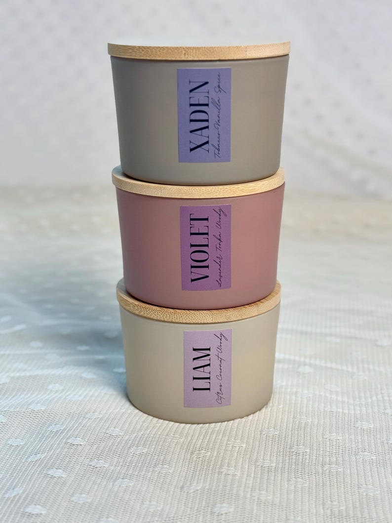 Może przedstawiać: Trzy ułożone w stos świece z drewnianymi pokrywkami. G&oacute;rna świeca jest szara z etykietą z napisem "XADEN Tobacco Vanilla Spice". Środkowa świeca jest r&oacute;żowa z etykietą z napisem "VIOLET Lavender Tonka Woody". Dolna świeca jest beżowa z etykietą z napisem "LIAM Citrus Coconut Woody".