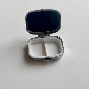 K&ouml;nnte beinhalten: Ge&ouml;ffnete, silberfarbene Pillendose mit wei&szlig;em Innenraum, der in zwei F&auml;cher unterteilt ist. Der Deckel ist ge&ouml;ffnet und zeigt eine dunkle, reflektierende Oberfl&auml;che. Die Pillendose ist rechteckig mit abgerundeten Kanten und einem Klappdeckel.
