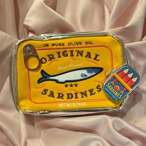 May include: A yellow sardine-themed zippered pouch with a silver trim. The pouch features a sardine illustration and the words "Original Sardines" and "Net Wt 3.75 oz". A small sardine tin is also visible.